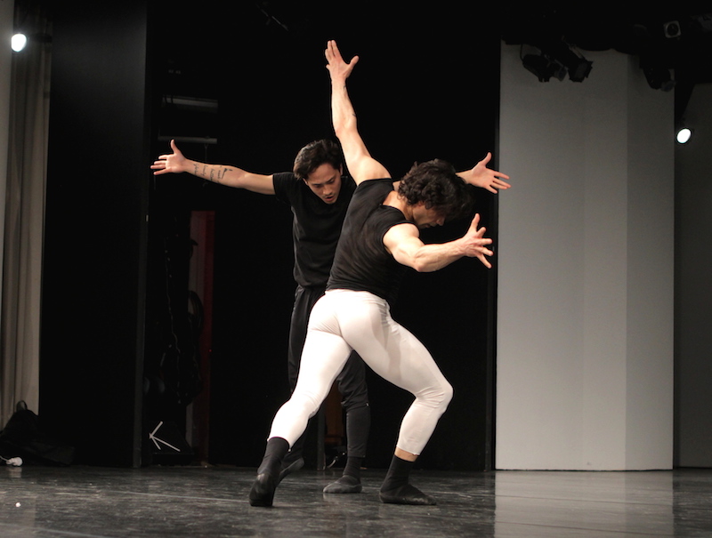 Two men open their arms and lunge toward each other. They wear black and white t-shirts and ballet tights.